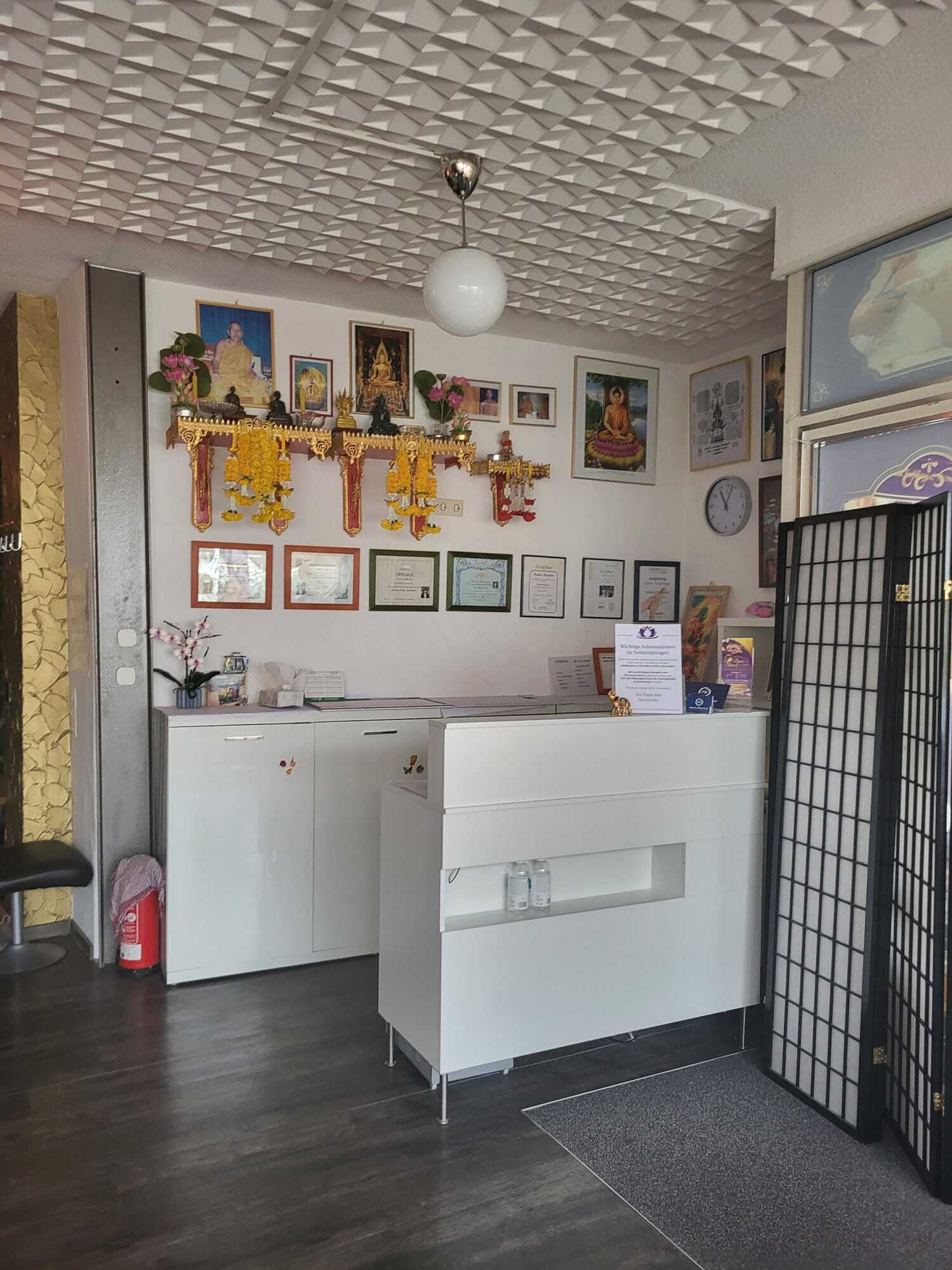 Empfangsbereich eines Studios oder Salons mit weißem Tresen, dekorativen buddhistischen Bildern und Figuren an der Wand, Urkunden und Zertifikaten sowie modernen Möbeln.