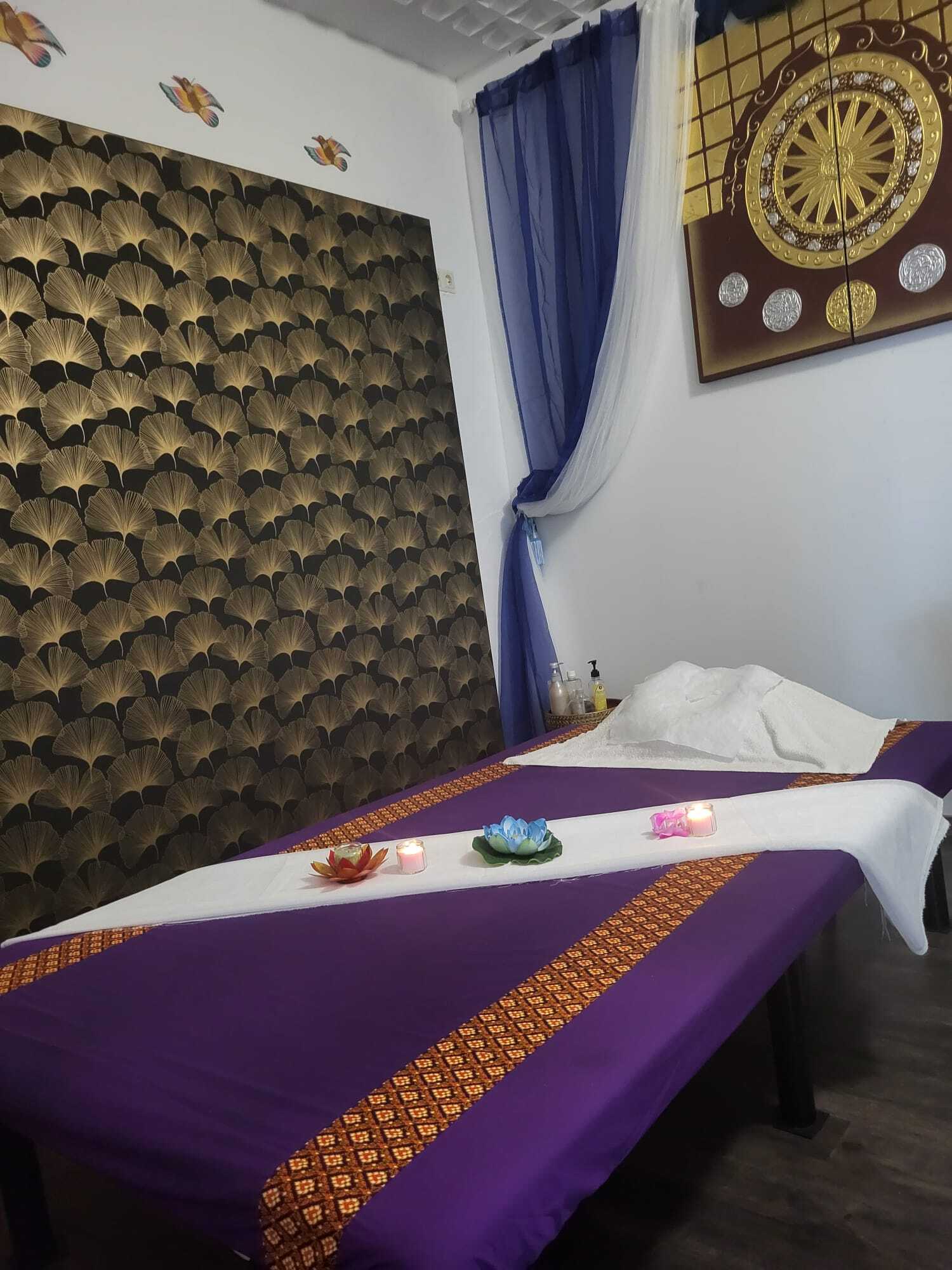 Stilvoll eingerichtetes Massagezimmer mit violettem Massagetisch, weißen Handtüchern, Blumen und Kerzen für Entspannung